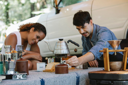 Positive young multiethnic couple in casual clothes switching on portable gas stove to boil water in coffee kettle while having picnic together in sunny nature