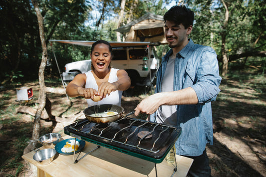 High angle of positive multiracial couple frying eggs on skillet using metal gas stove on wooden table near car and tent in forest