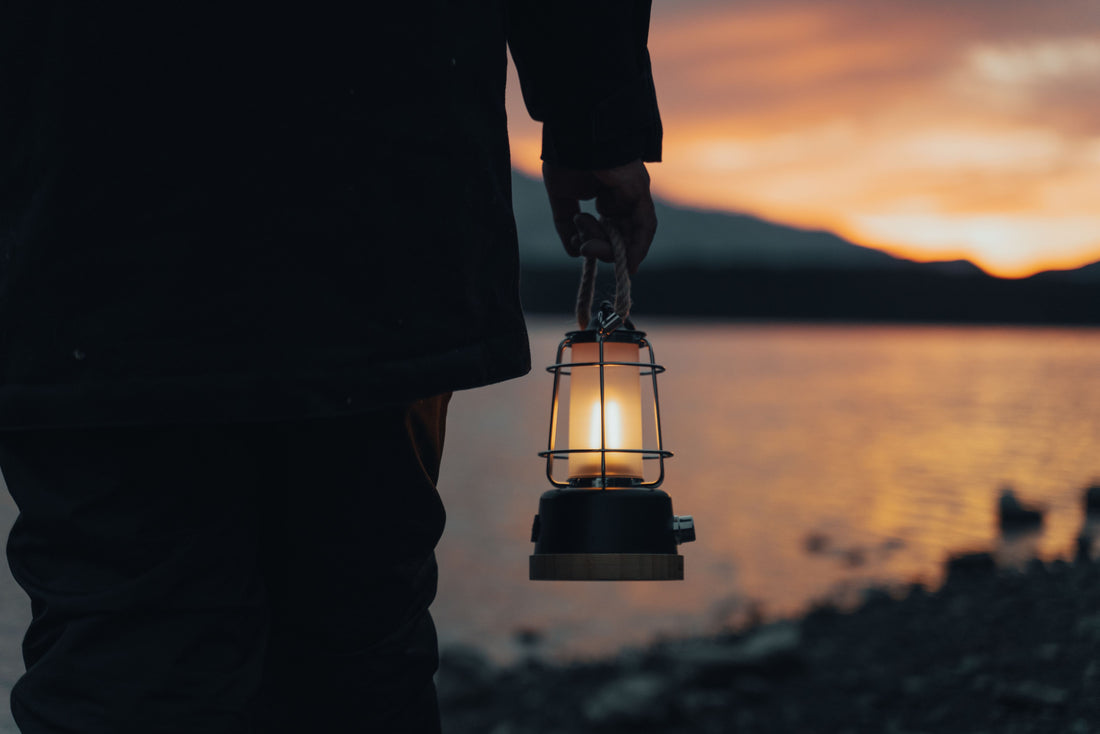 A person holding a glowing lantern beside a tranquil lake during sunset, creating a moody and calming scene.